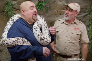 Gabriel Iglesias with a snake wrapped around his torso, standing beside host Jay Brewer.